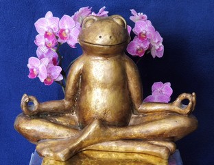 Eine goldene Froschfigur sitzt in Meditationshaltung vor einer Orchidee.